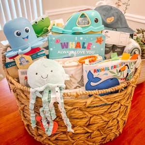 May include: A wicker basket filled with baby shower gifts, including a blue and green whale toy, a blue and white octopus toy, a blue and white jellyfish toy, a book titled "I Whale Always Love You", a blue and white animal friend book, and a pair of blue and white Nike socks.