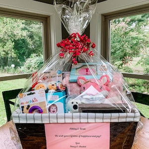 May include: A wooden gift basket filled with baby items, including a pink stuffed animal, a book, a rattle, and a teething ring. The basket is wrapped in clear cellophane and tied with a red ribbon. A pink card with the text "Ashley & Brad, We wish you a lifetime of happiness and joy!! Love, Stacy & Anabel" is attached to the basket.