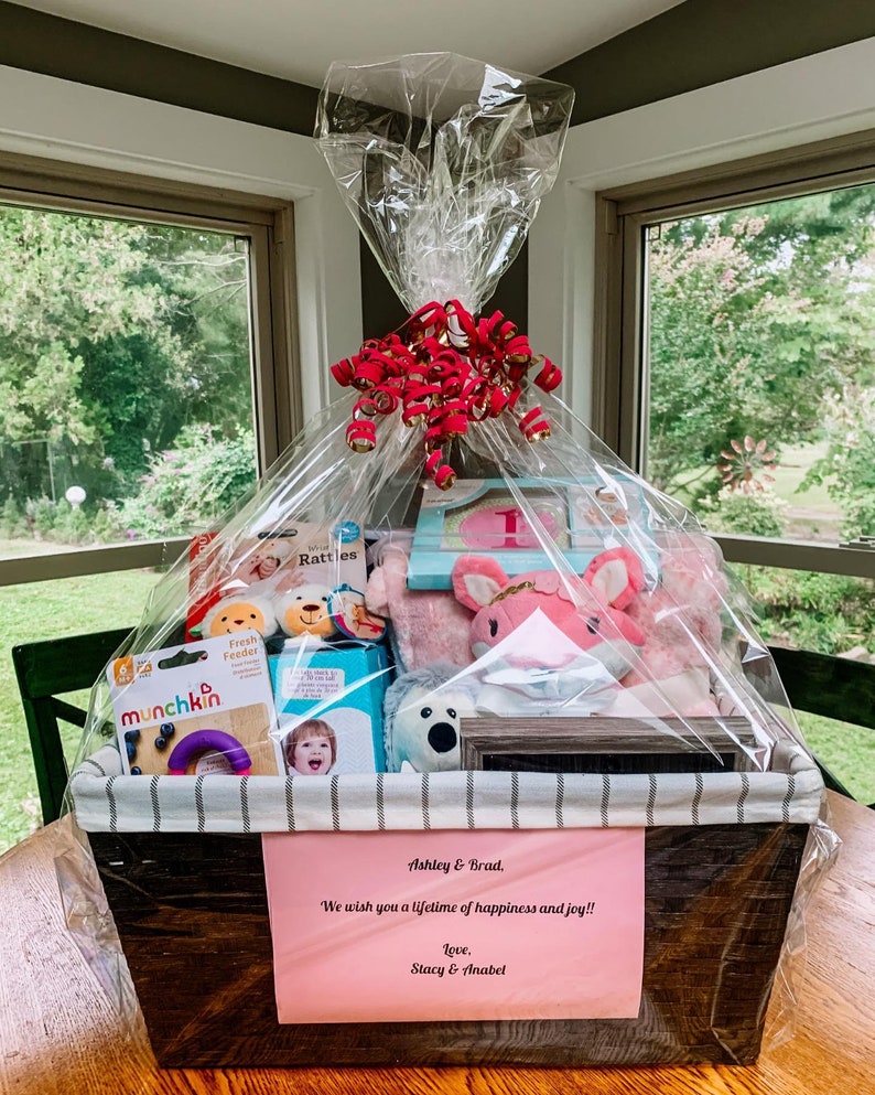 May include: A wooden gift basket filled with baby items, including a pink stuffed animal, a book, a rattle, and a teething ring. The basket is wrapped in clear cellophane and tied with a red ribbon. A pink card with the text "Ashley & Brad, We wish you a lifetime of happiness and joy!! Love, Stacy & Anabel" is attached to the basket.
