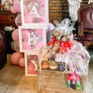 May include: A wooden crate filled with baby gifts, including bottles, a rattle, and baby shoes. The crate is decorated with a burlap bow and a pink ribbon. The crate is sitting on a carpet in front of a wall decorated with balloons and a baby block tower that spells "BABY".