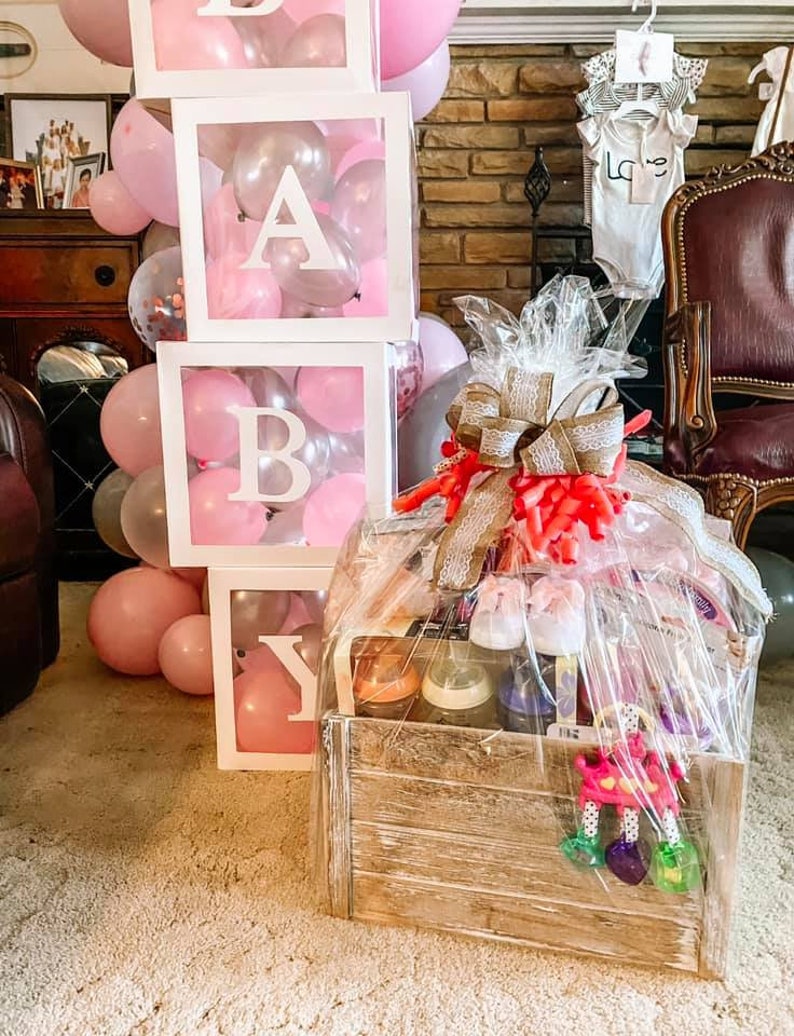 May include: A wooden crate filled with baby gifts, including bottles, a rattle, and baby shoes. The crate is decorated with a burlap bow and a pink ribbon. The crate is sitting on a carpet in front of a wall decorated with balloons and a baby block tower that spells "BABY".