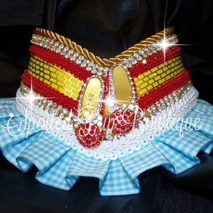May include: A decorative collar featuring a yellow brick pattern band, red trim, and gold rope edging. It is adorned with two miniature shoes covered in red rhinestones and clear crystals, finished with a ruffled light blue and white gingham fabric trim.