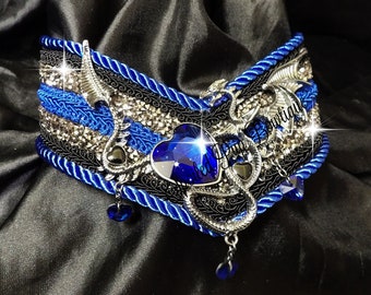 May include: A wide collar featuring layers of blue twisted rope trim, black decorative braid, and rows of silver rhinestones. A central heart-shaped blue gemstone is flanked by silver dragon figures, with small blue heart-shaped charms hanging below.
