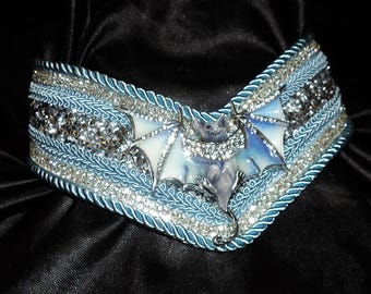 May include: A V-shaped collar featuring a central bat figure with iridescent wings, surrounded by layers of light blue braided trim, silver metallic cord, and rows of sparkling clear crystals set against a dark, textured fabric base.