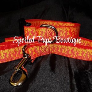 May include: A red and yellow patterned dog leash with a silver clasp and a D-ring. The leash has a flame-like design and the text "Spoiled Pups Boutique". The leash is designed for pets.