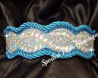 May include: A turquoise dog collar with a central band of iridescent rhinestones and smaller clear crystals. The collar is trimmed with a twisted rope design and the text "Spoiled Pups Boutique".