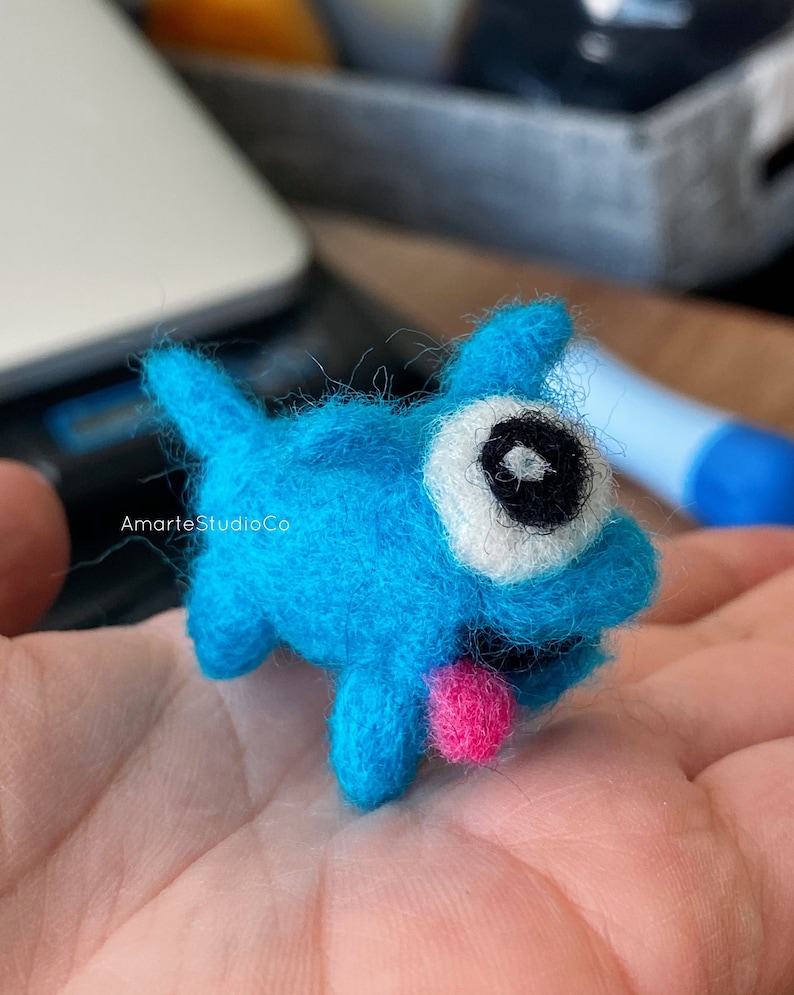 K&ouml;nnte beinhalten: Ein kleines, blaues Filzspielzeug mit einem gro&szlig;en Auge und einer herausgestreckten rosa Zunge. Das Spielzeug sitzt auf der Hand einer Person.