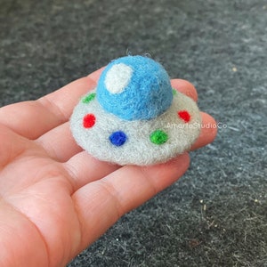 K&ouml;nnte beinhalten: Ein kleines Filz-UFO mit einer blauen Kuppel und einem wei&szlig;en Kreis oben. Das UFO hat eine graue Basis mit roten, blauen, gr&uuml;nen und wei&szlig;en Punkten.