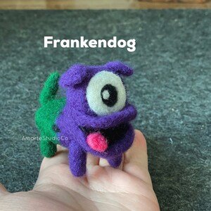 K&ouml;nnte beinhalten: Ein lila Filzspielzeug-Hund mit einem gr&uuml;nen Filzschwanz, einem gro&szlig;en wei&szlig;en Auge mit schwarzer Pupille und einer herausgestreckten rosa Zunge. Das Spielzeug ist mit "Frankendog" beschriftet.