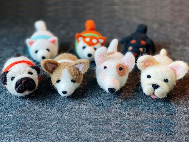 May include: A collection of eight felt animal figurines, each depicting a different breed of dog. The dogs are arranged in a row on a gray surface. The dogs are all white with different colored markings. The dogs are all looking at the camera.