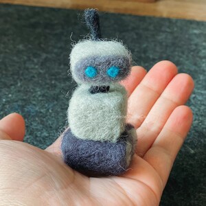 K&ouml;nnte beinhalten: Ein kleiner, grauer und schwarzer Filzroboter mit blauen Augen. Der Roboter besteht aus drei &uuml;bereinander gestapelten W&uuml;rfeln und hat eine schwarze Antenne oben drauf.
