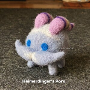 K&ouml;nnte beinhalten: Ein blauer und wei&szlig;er Filz-Poro mit violetten H&ouml;rnern und einem wei&szlig;en Schnurrbart. Der Poro steht auf einer grauen Oberfl&auml;che. "Heimerdinger's Poro" steht unter dem Poro.