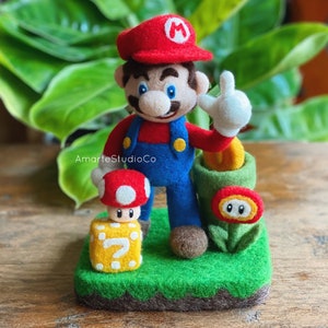 May include: A felted Mario figurine wearing a red cap and blue overalls, with a raised hand. A red and white mushroom, a question mark block, a green pipe, and a fire flower are also present on a green base. The text "AmateStudioCo" is visible.