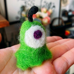 K&ouml;nnte beinhalten: Ein gr&uuml;nes Filztier mit einem einzigen wei&szlig;en Auge und einer violetten Pupille. Das Tier hat eine schwarze Filzantenne.