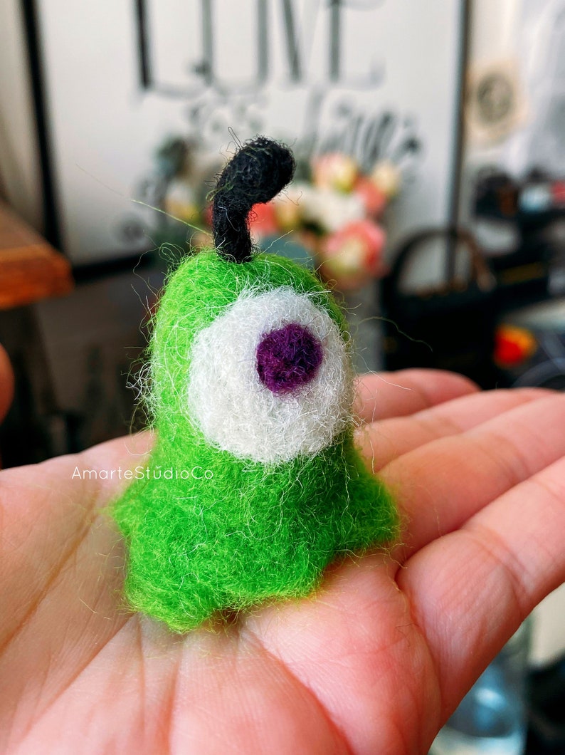 K&ouml;nnte beinhalten: Ein gr&uuml;nes Filztier mit einem einzigen wei&szlig;en Auge und einer violetten Pupille. Das Tier hat eine schwarze Filzantenne.