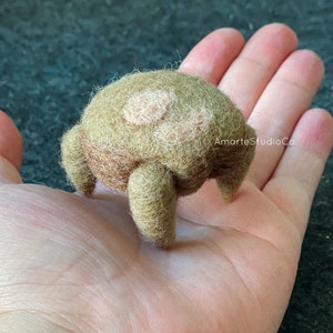 K&ouml;nnte beinhalten: Ein kleines, braunes und gr&uuml;nes Filztier mit zwei hellbraunen Flecken auf dem R&uuml;cken. Es hat vier Beine und steht auf einer Hand.
