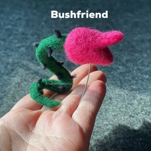 K&ouml;nnte beinhalten: Eine Filzskulptur einer rosa Blume mit einem gr&uuml;nen Stiel und schwarzen Dornen. Die Skulptur wird in einer Hand gehalten und der Text "Bushfriend" ist &uuml;ber der Skulptur sichtbar.