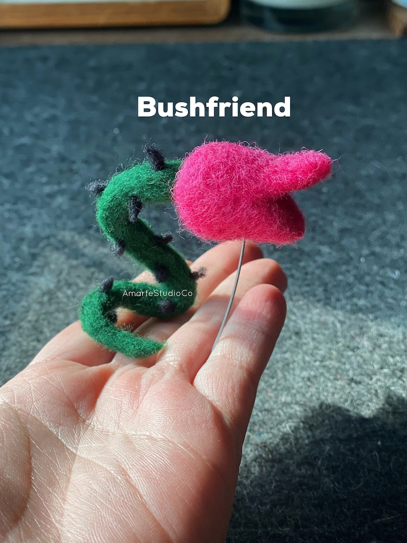 K&ouml;nnte beinhalten: Eine Filzskulptur einer rosa Blume mit einem gr&uuml;nen Stiel und schwarzen Dornen. Die Skulptur wird in einer Hand gehalten und der Text "Bushfriend" ist &uuml;ber der Skulptur sichtbar.