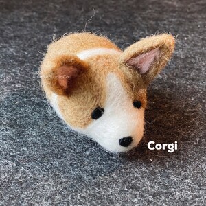 May include: A small, brown and white felt corgi figurine with black eyes and a black nose. The figurine is lying on its side with its head turned to the right. The text "Corgi" is written in white letters on a black background below the figurine.