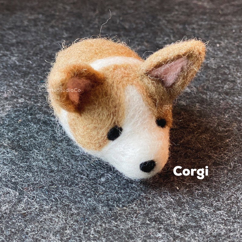 May include: A small, brown and white felt corgi figurine with black eyes and a black nose. The figurine is lying on its side with its head turned to the right. The text "Corgi" is written in white letters on a black background below the figurine.