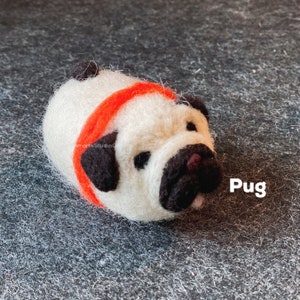 May include: A white felt pug with black ears and a black nose, wearing a red felt headband. The pug is lying on its side with its tongue sticking out. The word "Pug" is written in black text on a gray background.