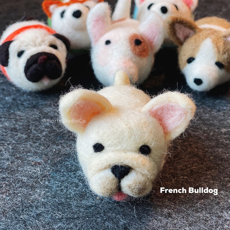 May include: A close-up of a white felt French Bulldog figurine with black eyes and a pink nose. The figurine is lying on its side with its tongue sticking out. The text "French Bulldog" is written below the figurine.