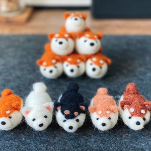May include: A collection of handmade felt Shiba Inu dog figurines. The figurines are white, orange, and black, with small black eyes and noses. Some are stacked in a pyramid shape, while others are arranged in a row.