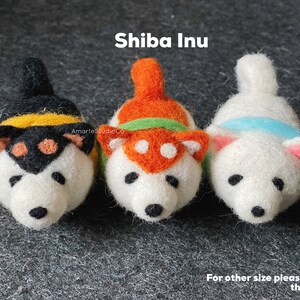 May include: Three felt Shiba Inu dog figurines. The dogs are white with different colored accents. The first dog has black and yellow accents, the second dog has orange and green accents, and the third dog has blue accents. The dogs are all looking at the camera.