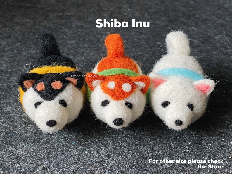 May include: Three felt Shiba Inu dog figurines. The dogs are white with different colored accents. The first dog has black and yellow accents, the second dog has orange and green accents, and the third dog has blue accents. The dogs are all looking at the camera.