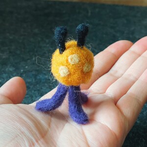 K&ouml;nnte beinhalten: Ein kleines, handgefilztes Wesen mit einem gelben K&ouml;rper, lila Beinen und schwarzen Antennen. Das Wesen hat drei wei&szlig;e Punkte auf seinem K&ouml;rper.
