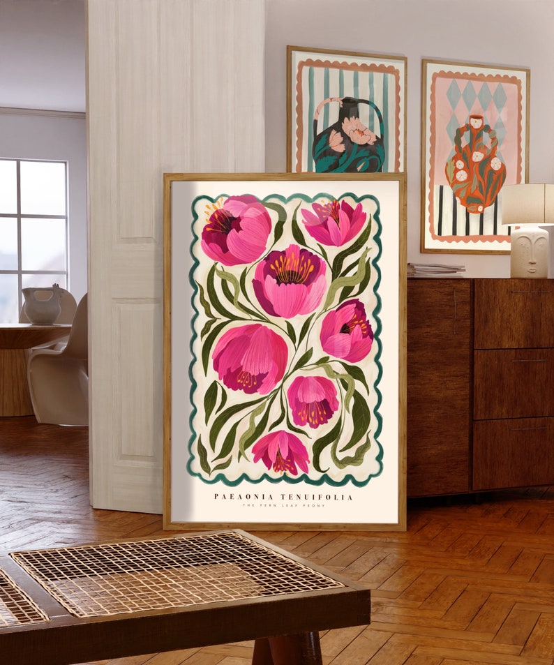 May include: A framed botanical illustration features pink Paeonia tenuifolia, also known as the fern-leaf peony, depicted in a stylized manner against a cream background with teal scalloped border.  The artwork is presented in a light wood frame and displayed in a mid-century modern interior setting, alongside two additional framed prints featuring abstract vase designs in a retro color palette. The overall aesthetic is vintage-inspired, with a focus on botanical art and mid-century modern design elements.
