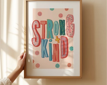 May include: A framed print with a pink background and the words "Strong + Kind" in colourful, patterned letters. The letters are surrounded by small, white smiley faces.
