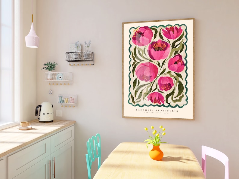 May include: Pink floral poster print featuring Paeonia tenuifolia, the fern-leaf peony, in a vintage style. The artwork showcases a cluster of vibrant pink flowers with green foliage, set against a cream background within a decorative border. The poster's dimensions are not specified, but it's displayed in a light-colored room with teal and pink chairs, a light-colored kitchen counter, and a small vase of yellow flowers.