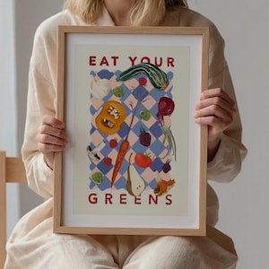 May include: A framed print with a light blue and pink checkered background. The print features a variety of fruits and vegetables, including a pear, a carrot, a tomato, a squash, and a mushroom. The text "EAT YOUR GREENS" is printed in red at the top and bottom of the print.