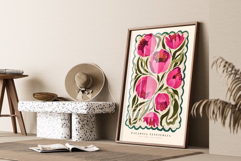 May include: Framed botanical illustration featuring pink Paeonia tenuifolia flowers on a cream background with dark green accents. The print has a decorative scalloped border.  The artwork's title, 'Paeonia Tenuifolia,' is visible at the bottom. The print is displayed against a neutral backdrop, leaning against a terrazzo side table with a hat and magazines nearby.