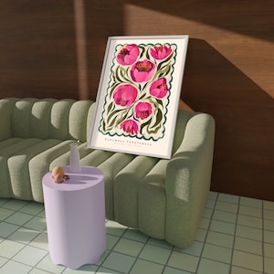 May include: A framed botanical illustration featuring pink Paeonia tenuifolia flowers on a cream background with green foliage. The artwork has a dark teal border. The text 'PAEONIA TENUIFOLIA' is printed at the bottom. The print is displayed on a light green sofa against a brown wall. A small light purple side table with decorative items sits in front of the sofa.