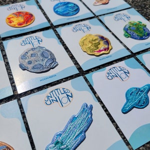 May include: A set of nine embroidered iron-on patches featuring planets and celestial bodies. The patches are on white cards with the text "Untitled Union" and the name of the celestial body. The patches are blue, green, yellow, and orange.
