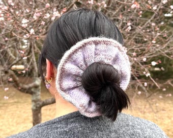 May include: A knitted scrunchie in shades of lavender, gray, and brown, worn in the hair. The scrunchie is wrapped around a dark bun. The person is wearing a gray sweater. The background features a tree with pink blossoms.