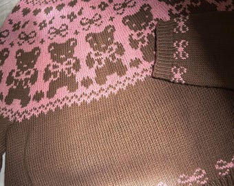 May include: Brown and pink knit sweater featuring a teddy bear and bow pattern. The sweater has a crew neck and long sleeves. The bottom and cuffs of the sleeves have a bow pattern. The sweater is made of a soft, knitted material.
