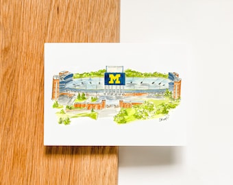 May include: A watercolor illustration of Michigan Stadium, also known as "The Big House", with the iconic "M" on the scoreboard. The stadium is surrounded by green trees and a brick walkway.