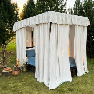 May include: A canopy bed frame with white linen curtains and a blue bedspread. The bed is set up outdoors in a grassy area with trees in the background. The text "Dusty Linen" is visible at the bottom of the image.