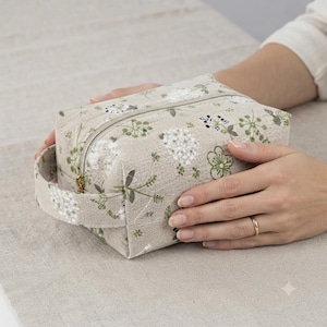 May include: A rectangular beige fabric pouch featuring a repeating pattern of white and green botanical floral illustrations. The pouch includes a top zip closure and a side handle made from the same patterned material, with visible stitching.