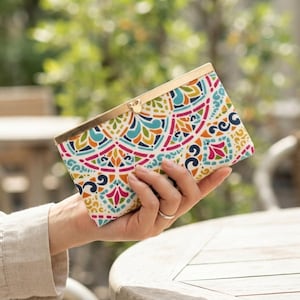 May include: A rectangular clutch wallet featuring a vibrant, multi-coloured geometric pattern in shades of teal, magenta, yellow, and orange. The wallet has a gold-toned metal frame with a small, circular clasp closure at the top centre.