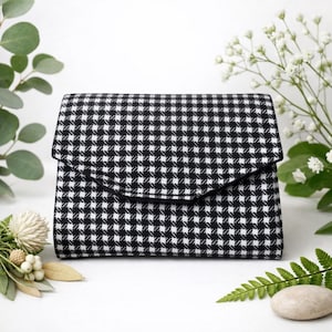 Peut inclure: Une pochette rectangulaire de style enveloppe arborant un motif pied-de-poule classique noir et blanc. Le sac présente un rabat structuré qui recouvre l'avant, mettant en valeur un motif textile à petits carreaux répétitif et uniforme.
