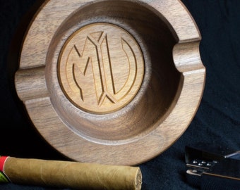 May include: A wooden cigar ashtray with a carved monogram "MU" in the center. A cigar and a silver lighter are also pictured on a black surface.