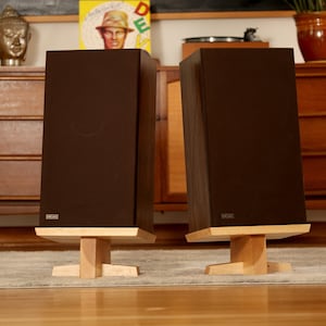 Puede incluir: Dos gabinetes de altavoces de madera marrón oscuro con rejillas de altavoces negras y una pequeña etiqueta rectangular en la parte delantera. Los altavoces están sobre soportes de madera con un acabado de madera clara. Los altavoces están delante de un armario de madera con cajones.