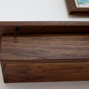May include: A dark brown wooden shelf with a black metal hook attached to the top. The shelf is mounted to a white wall.