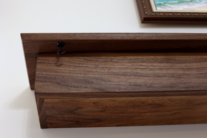 May include: A dark brown wooden shelf with a black metal hook attached to the top. The shelf is mounted to a white wall.