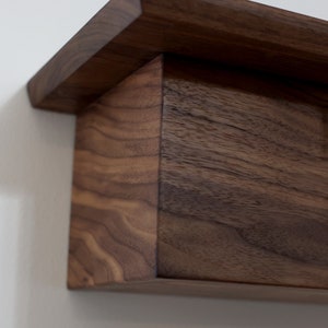 May include: A dark brown wooden shelf with a simple, rectangular design. The shelf has a slightly angled top edge and is made of solid wood with visible grain.