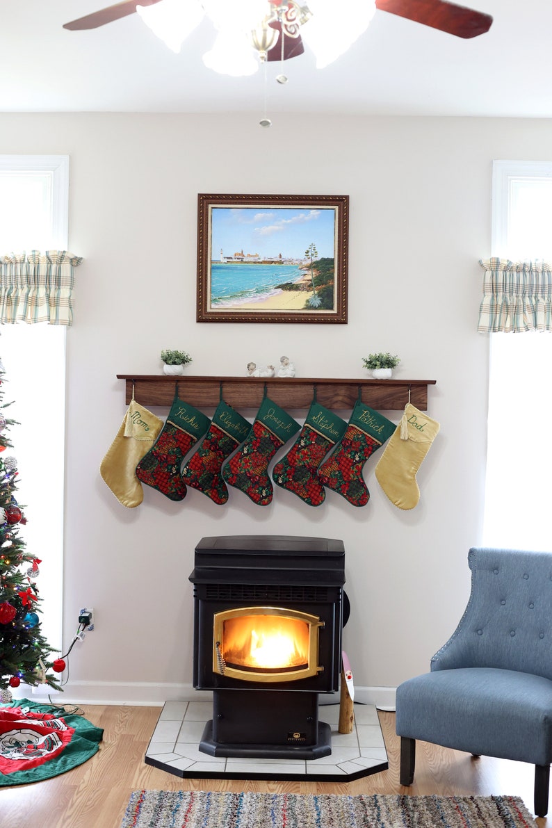 May include: A wood mantel with seven stockings hanging from it. The stockings are green with red and gold accents and have names embroidered on them. There is a wood-burning stove with a fire in it below the mantel. A blue armchair is to the right of the stove.